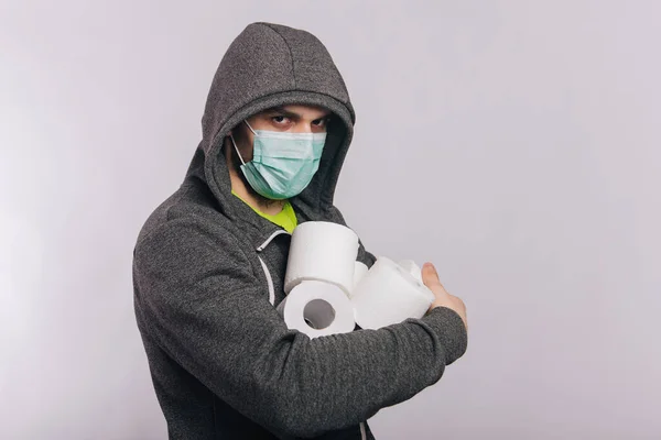 Un gars en sweat-shirt gris et avec des rouleaux de papier toilette est debout sur un fond blanc. L'excitation pendant la quarantaine pour le coronavirus. Rotavirus chez un homme portant un masque médical . — Photo