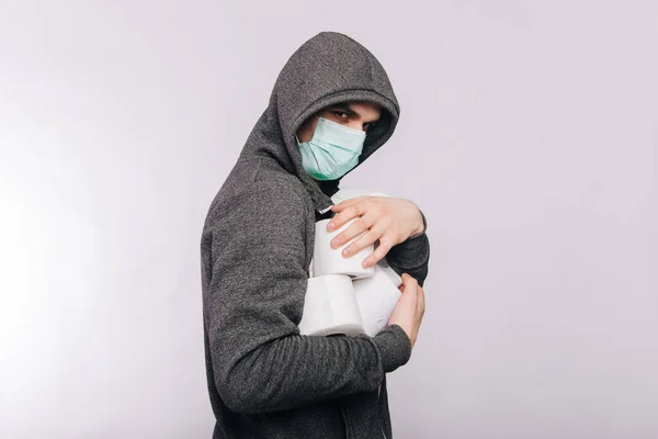 Un gars en sweat-shirt gris et avec des rouleaux de papier toilette est debout sur un fond blanc. L'excitation pendant la quarantaine pour le coronavirus. Rotavirus chez un homme portant un masque médical . — Photo