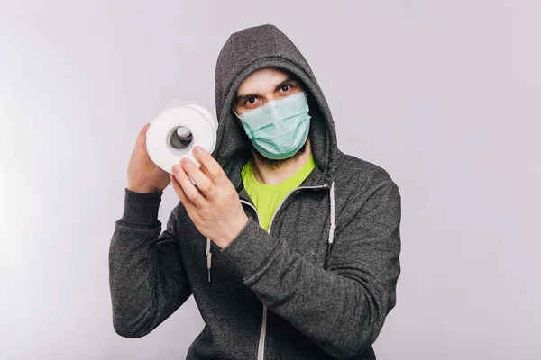 Ένας τύπος με γκρι μπλούζα και ρολό χαρτί τουαλέτας στέκεται σε λευκό φόντο. Ο ενθουσιασμός κατά τη διάρκεια της καραντίνας για τον κορωναϊό. Ροταϊοί σε έναν άντρα με ιατρική μάσκα.. — Φωτογραφία Αρχείου