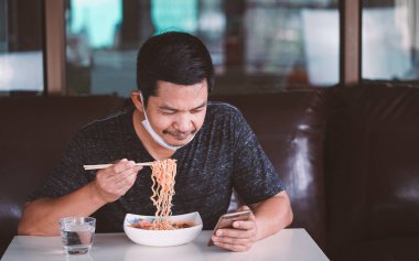 Maskeli bir adam erişte yiyor ve evde akıllı bir telefon tutuyor. 2019-ncov virüsünü veya Covid-19 Concept.16: 9 tarzını korumak için evde kalın.