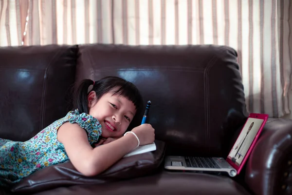 Happy little child girl lying on sofa with learning by african teacher on laptop and writing a book at home,Social distance during quarantine, Online education concept