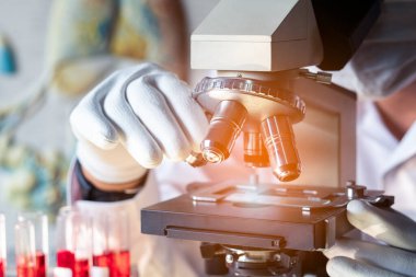 Bilim adamı ya da doktor mikroskop altında kan örneklerine bakarken tıbbi koruyucu maske takıyor.