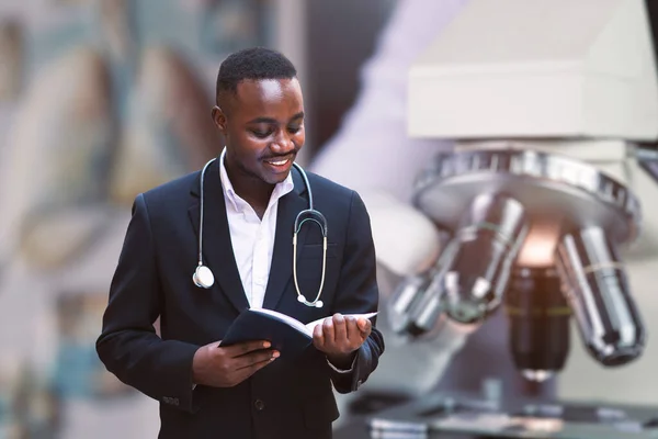 African doctor with stethoscope wearing classic suit reading a book  near modern clinic hospital building