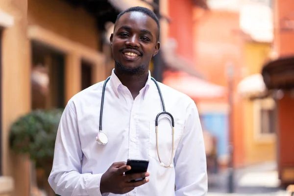 African doctor with stethoscope holding a smartphone near modern clinic hospital building