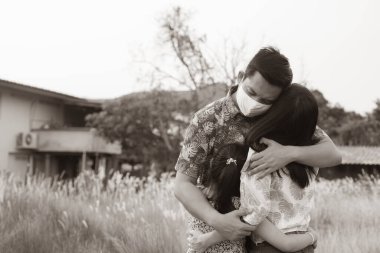 Baba yüz maskesi takıyor. Ailesine sarılıyor. Sevgiyle geri çekiliyor. Covid-19 pandemik konsepti boyunca sosyal mesafe evde kalır. Siyah-beyaz tarzı.