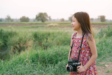 Küçük kız elinde orta formatlı film kamerası ve yeşil alan arka planlı günbatımı manzarasının fotoğrafını çekiyor.