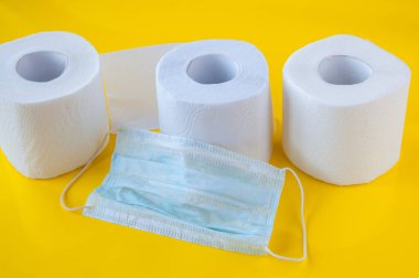 Üç rulo beyaz tuvalet kağıdı ve sarı arka planda tıbbi maske.
