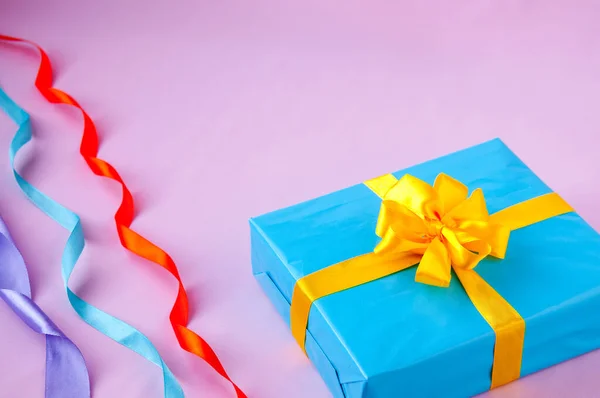 Gift in a blue box with an yellow bow and multi-colored ribbon on a pink background