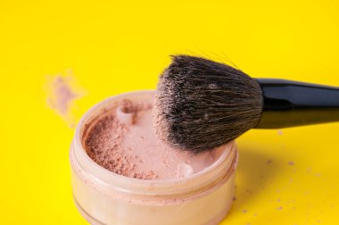 Açık bir toz kavanozu ve sarı arka planda büyük bir kozmetik fırçası.