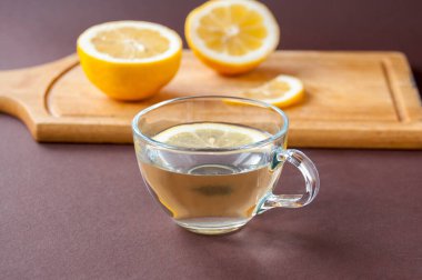 Tahta kesim tahtasının üzerinde kesilmiş bir limon ve kahverengi arka planda limon olan şeffaf bir kupa var.