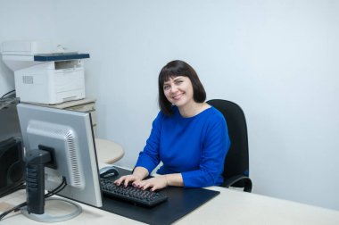 Ofis, mavi elbiseli esmer kadın masasına gülümsüyor.
