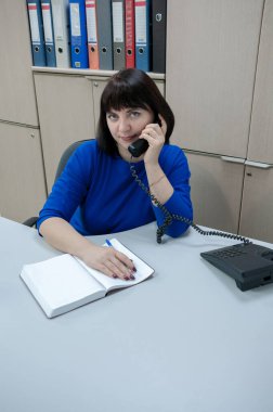 Mavi elbiseli bir kadın telefonda ofiste bir müşteriyle konuşuyor.