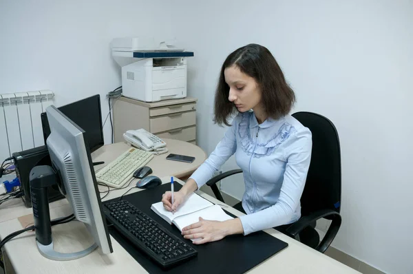 Esmer kız bilgisayarın ofisindeki bir deftere notlar alıyor.