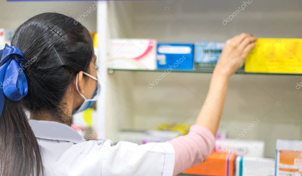 Female pharmacist taking a medicine from the shelf, wearing  lab coat during work in a modern drugstore with various pharmaceutical products. 