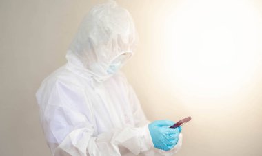 Doktor, Coronavirus 'u (COVID19) korumak için PPE giysisi giymekte ve Corona / covid-19 virüsü enfekte olmuş hastasının laboratuarına başvuru telefonu raporuyla bakmaktadır. Virüsle savaşmak için Covid-19 karantinası.