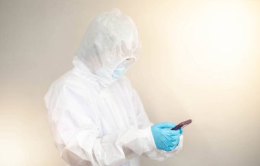 Doktor, Coronavirus 'u (COVID19) korumak için PPE giysisi giymekte ve Corona / covid-19 virüsü enfekte olmuş hastasının laboratuarına başvuru telefonu raporuyla bakmaktadır. Virüsle savaşmak için Covid-19 karantinası.