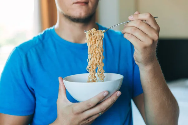 Young handsome man in a blue T-shirt eating instant noodles from a white plate for lunch at home, concept of cheap food or food delivery, selective focus, close-up