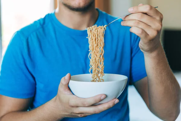 Young handsome man in a blue T-shirt eating instant noodles from a white plate for lunch at home, concept of cheap food or food delivery, selective focus, close-up