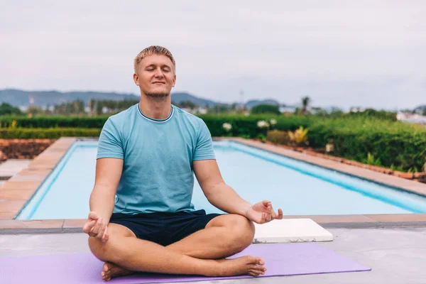 Handsome young blond man plays sports outdoors by the pool in tropics during quarantine, guy meditates in a lotus position, holds his hands in mudra, the concept of independent training and fitness