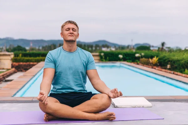 Handsome young blond man plays sports outdoors by the pool in tropics during quarantine, guy meditates in a lotus position, holds his hands in mudra, the concept of independent training and fitness