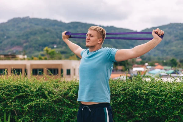 Handsome young blond man plays sports alone at home outdoors in the tropics during quarantine, guy is training with elastic resistance band, the concept of independent training and fitness, close-up