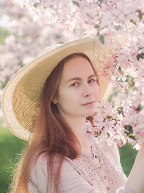 Çiçekli elma bahçesi. Yakın plan portre. Hasır şapkalı ve pembe bluzlu beyaz bir kız. Ağaçlardaki çiçekler pembedir. Kızın uzun kızıl saçları rüzgarda dalgalanıyor. Gün batımı parlak fotoğraf