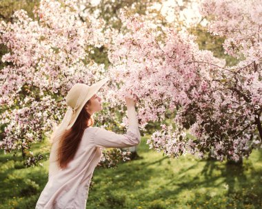 Çiçekli elma bahçesi. Hasır şapkalı ve pembe bluzlu beyaz bir kız. Ağaçlardaki çiçekler pembedir. Kızın uzun kızıl saçları rüzgarda dalgalanıyor. Gün batımı parlak fotoğraf