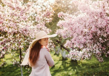 Çiçekli elma bahçesi. Hasır şapkalı ve pembe bluzlu beyaz bir kız. Ağaçlardaki çiçekler pembedir. Kızın uzun kızıl saçları rüzgarda dalgalanıyor. Gün batımı parlak fotoğraf