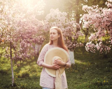Çiçekli elma bahçesi. Hasır şapkalı ve pembe bluzlu beyaz bir kız. Ağaçlardaki çiçekler pembedir. Kızın uzun kızıl saçları rüzgarda dalgalanıyor. Gün batımı parlak fotoğraf