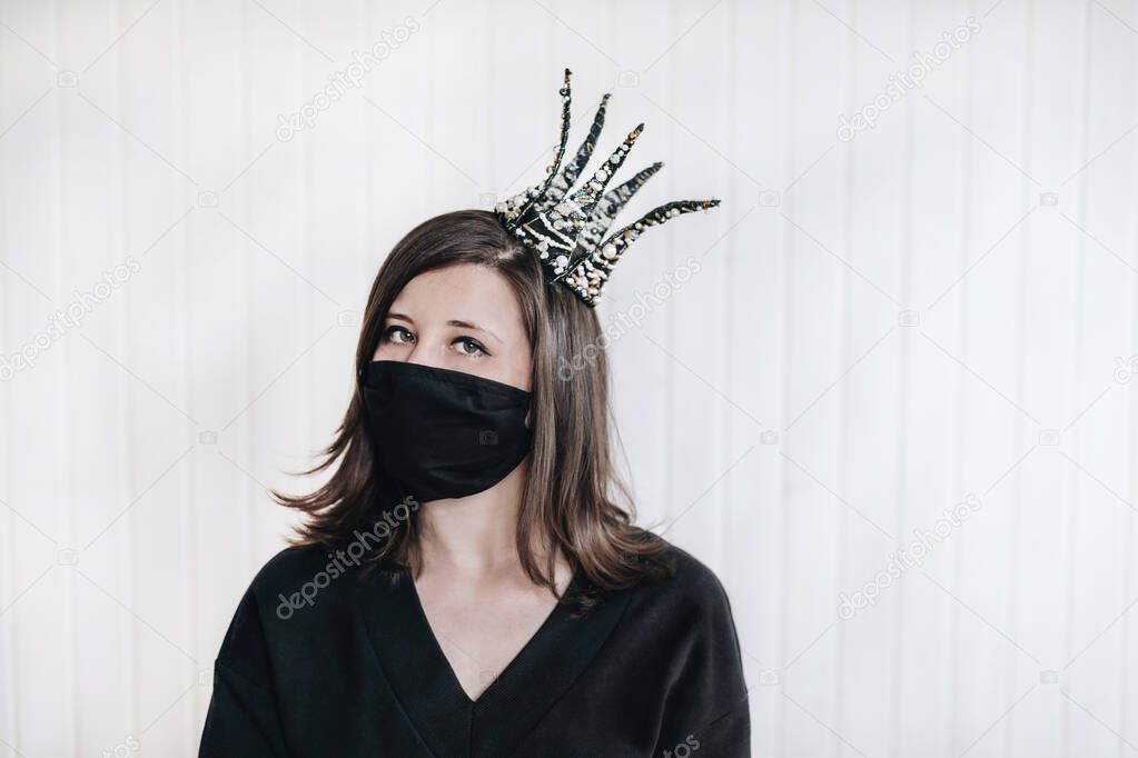 A girl on a white background wearing a medical mask and a crown on her head. Protection from coronavirus. The girl looks straight ahead. The background is white, the clothes are black. Black medical mask.