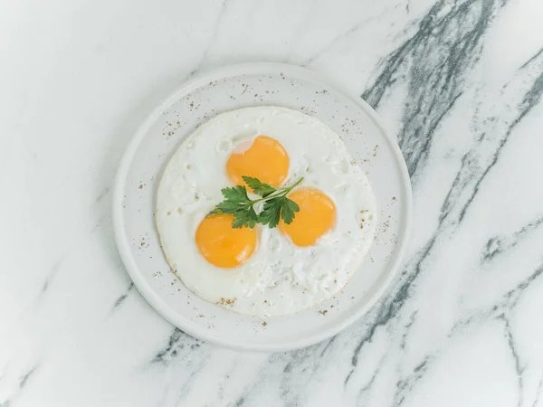 Breakfast is three eggs fried on a white plate on a white marble background. Decorated with parsley. Scrambled eggs with cheese. Photo for the menu.