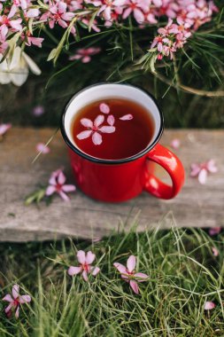 Sokakta çay partisi. Kırmızı bardaktaki çay yeşil çimlerin üzerindeki ahır tahtasının üzerinde duruyor. Pembe çiçekli dalların yanında. Kupada bir çiçek süzülür..