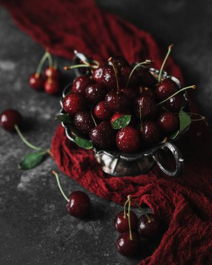 Eski bir kasede kırmızı bir kiraz uzun kırmızı bir peçetenin üzerinde duruyor. Yakınlarda böğürtlen var. Gri, karanlık bir arka planda hayat. Yaz havası..