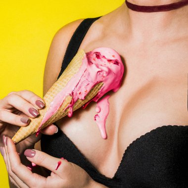 Dondurma kızın göğsünde eriyor. Arka plan sarı. Waffle külahında pembe dondurma. Giysiler siyah. Açık yaka. Boynunda kırmızı bir kurdele var. Yemek ve kadınların göğüsleri.