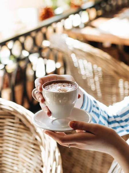 Morning at the cafe. A Cup of cappuccino with cinnamon. The girl collects foam with a spoon. The Cup is white. It\'s Sunny and warm outside. The atmosphere of an Italian restaurant or cafe. Wicker furniture in the background.