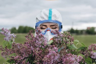 Covid-19 virüsünden koruyucu giysili kız. Yüzünde maske ve gözlük var. Bir insan bir buket leylağı örtüyor. Corona virüsü. Karantinaya alın. Salgın. Kendini soyutlama. Nokta. 