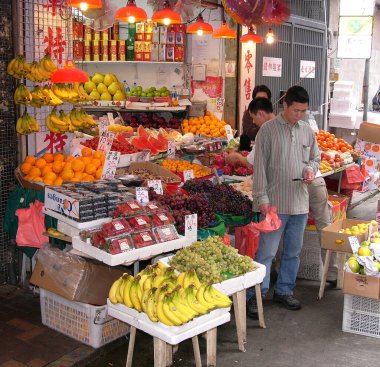 13 Mar 2012. Taze meyve tezgahı, Hong Kong. Sergide, bazıları plastik kaplarda ve diğerleri havaya açık birçok farklı meyve çeşidi yer alıyor. Ön planda cep telefonuna bakan bir adam var.