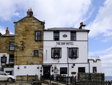 18 Ağustos 2018: The Bay Hotel, Robin Hoods Bay, North Yorkshire, İngiltere. Beyaz bina, kıyı şeridine karşı, mavi, bulutlu gökyüzüne karşı. Turistler giriş kapısının yakınındaki içeceklerden hoşlanırlar..