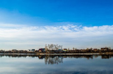 Ufuk mavi gökyüzünü bulutlarla ve gökyüzünü yansıtan bir nehirle evleri ayırıyor.