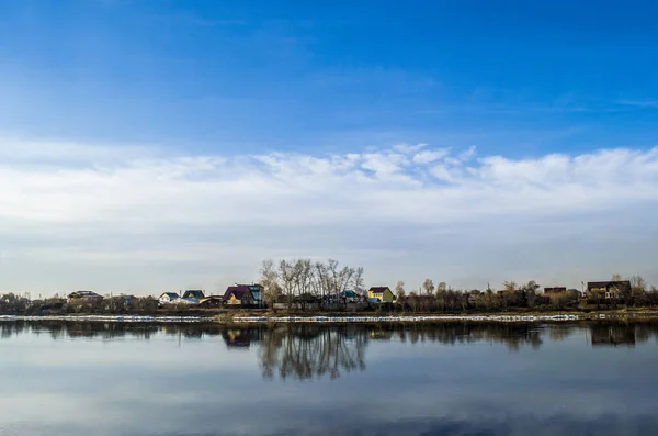 Ufuk mavi gökyüzünü bulutlarla ve gökyüzünü yansıtan bir nehirle evleri ayırıyor.
