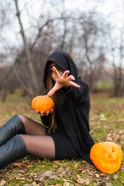 Bir kız, elinde balkabağıyla çimenlerin üzerinde oturuyor. Elinde yapraklar, elini yüzünün önünde bir kapüşon ve ormanın ortasında siyah bir manto ile tutuyor..