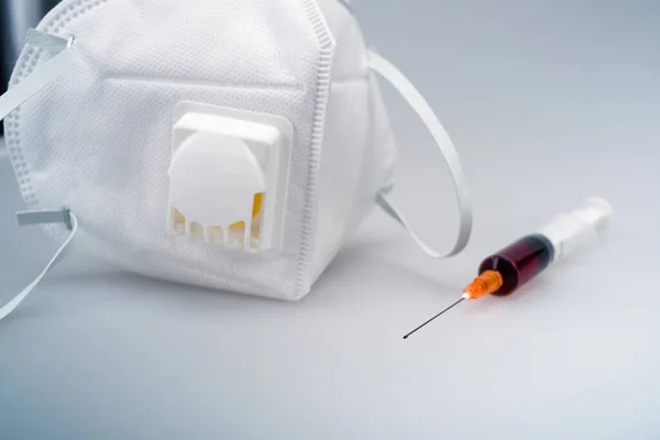 Havalandırmalı N95 koruyucu maske, anti-sis maskesi, kırmızı sıvı ilaç dolu bir şırınga, tıbbi ekipman. Mavi arka planda beyaz n95 maskesi ve plastik şırınga.