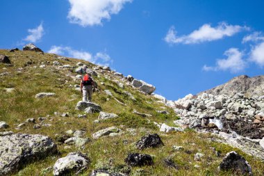 Bir adam kırmızı sırt çantalı bir turist bir kaya yamacına tırmanıyor. Bir turist dikkatlice yokuş yukarı yürür. Bir dağ deresi boyunca. Mavi gökyüzü ve beyaz bulutlar. Metin için alanı kopyala.