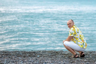 Limonlu tişörtlü bir adam turkuaz deniz kıyısında oturur ve çerçeveye bakar.