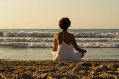 Afrika kökenli siyahi kadın beyaz bir elbise giyerek gün doğumunda kumsalda düşünüyor. meditasyon