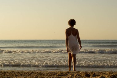 Sahilde, beyaz bir elbise giymiş Afro-Amerikalı siyahi bir kadın. Denize doğru yürüyorum