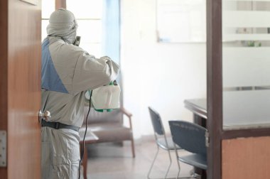 COVID-19 'u önlemek için ofisin dezenfekte edilmesi, ofiste dezenfekte edilmiş beyaz tehlikeli madde giysisi giyen kişi koruyucu maske takıyor ve Coronavirus püskürtüyor..