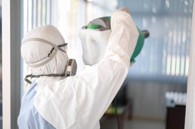 COVID-19 'u önlemek için ofisin dezenfekte edilmesi, ofiste dezenfekte edilmiş beyaz tehlikeli madde giysisi giyen kişi koruyucu maske takıyor ve Coronavirus püskürtüyor..