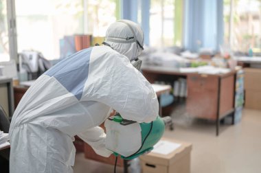 COVID-19 'u önlemek için ofisin dezenfekte edilmesi, ofiste dezenfekte edilmiş beyaz tehlikeli madde giysisi giyen kişi koruyucu maske takıyor ve Coronavirus püskürtüyor..