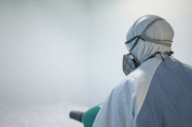 COVID-19 'u önlemek için ofisin dezenfekte edilmesi, ofiste dezenfekte edilmiş beyaz tehlikeli madde giysisi giyen kişi koruyucu maske takıyor ve Coronavirus püskürtüyor..